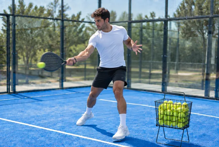 Een padeltrainer slaat een bal, met de mand met ballen naast zich op de baan